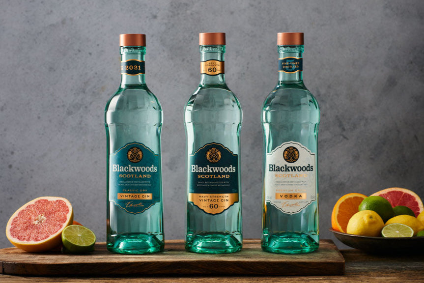 Three bottles of gin arranged on a wooden surface with citrus fruit beside them, against a grey background.